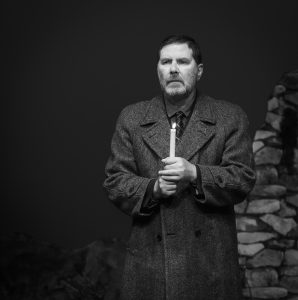 Black and white image of Bill Livingston played by Garry Kennedy. Bill stands alone, candle in hand as part of the candlelight vigil. He stares out to the audience with a blank, lost expression.