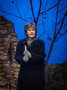 Woman Number 1 played by Jean Byrne looks off into the audience, lost in her thoughts, lit candle in hand as the candlelight vigil is held in the hills around Lockerbie.