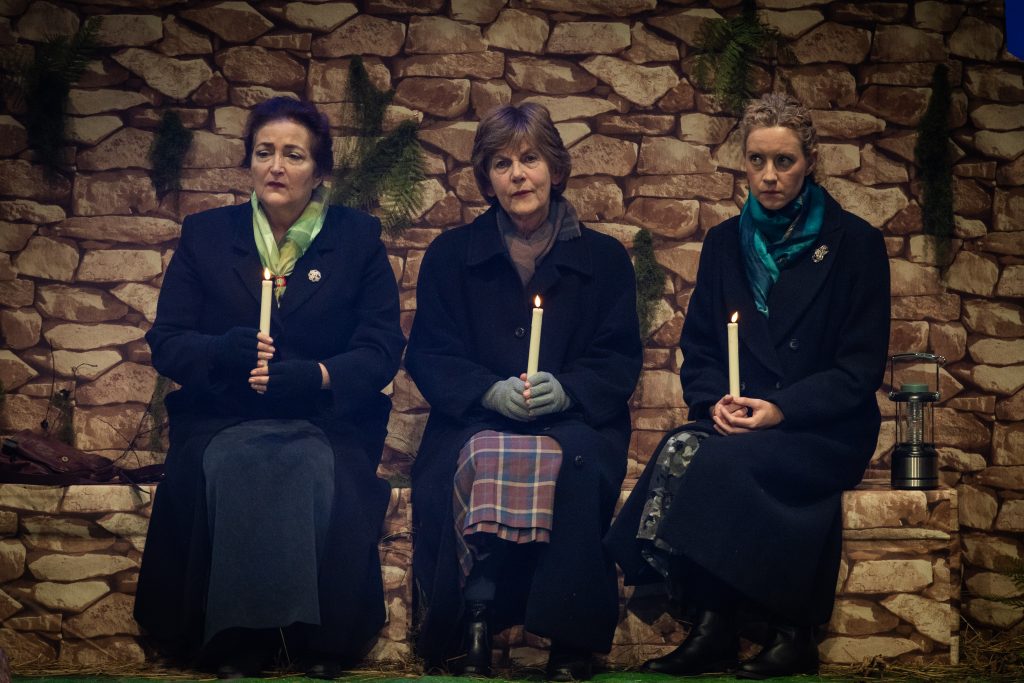 Three actors sit on stage the setting the hills of Lockerbie, Scotland. They are dressed for the cold Scottish weather and stare out into the audience with a look of sorrow. The actors are playing characters from the play "The Women of Lockerbie" by Deborah Brevoort. The image is in colour and shows a drystone dyke in the background which the actors sit with their backs to.