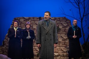 The candlelit vigil has started in the hills around Lockerbie and Olive, Woman number 1 and Number 2 stand with Bill. Bill in the foreground looks defeated with his struggles to keep Madeline grounded, stares off to where Madeline has again disappeared into the evening to wander the hills.