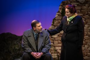 Olive consoles Bill Livingston by placing a hand on his shoulder. Bill is sitting on a rock as Olive stands by his side. They are set in the Lockerbie hills. In the background the colours depict a late evening with the sun having not long set.