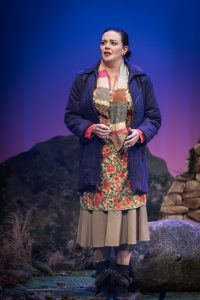 Hattie, the warehouse cleaner, stands in the hills of Lockerbie after telling the others how her plan to obtain the clothing of the missing has failed. She looks off into the distance with a look of sorrow as the colours in the background; blues, purples and pink, signify the sunset.