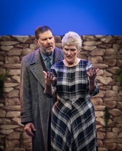 Madeline & Bill Livingston standing in the hills of Lockerbie. Madeline has cut herself after searching a warehouse for remnants of her son's belongings. She stands in front of Bill facing the audience, her hands raised, and blood on her face.